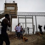 Valla fronteriza de Ceuta vigilada por patrullas policiales frente a un grupo masivo de personas en el lado de Marruecos.