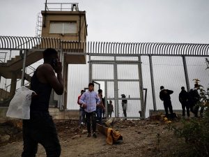 Valla fronteriza de Ceuta vigilada por patrullas policiales frente a un grupo masivo de personas en el lado de Marruecos.